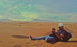 A person is sitting on a sandy desert with mountains in the distance, carefully examining a camera. They wear casual clothing and a cap, with a backpack beside them. The sky is partly cloudy, creating a dramatic effect over the arid landscape.