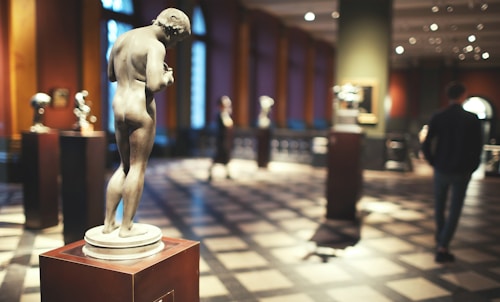 A classical-style sculpture is prominently displayed on a pedestal in an art gallery. The room has a checkered floor, warm lighting, and framed artworks hanging on the walls. A person is seen in the distance, walking past other displayed sculptures.