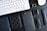 A collection of electronic and personal items is arranged neatly on a black surface. The items include a white keyboard and mouse, a smartwatch with two interchangeable black bands, a smartphone with a black case, a black embossed wallet, a black book or notebook, and a small square accessory with a metallic finish.