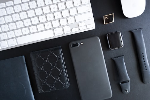 A collection of electronic and personal items is arranged neatly on a black surface. The items include a white keyboard and mouse, a smartwatch with two interchangeable black bands, a smartphone with a black case, a black embossed wallet, a black book or notebook, and a small square accessory with a metallic finish.