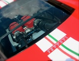 A close-up view of a car engine visible through a glass panel. The engine features prominent red components and is surrounded by intricate mechanical parts. The exterior surrounding the glass is a bright red with white and green racing stripes.