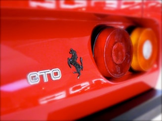 A close-up of the rear portion of a red sports car, featuring a black prancing horse emblem, the letters GTO, and rear taillights.