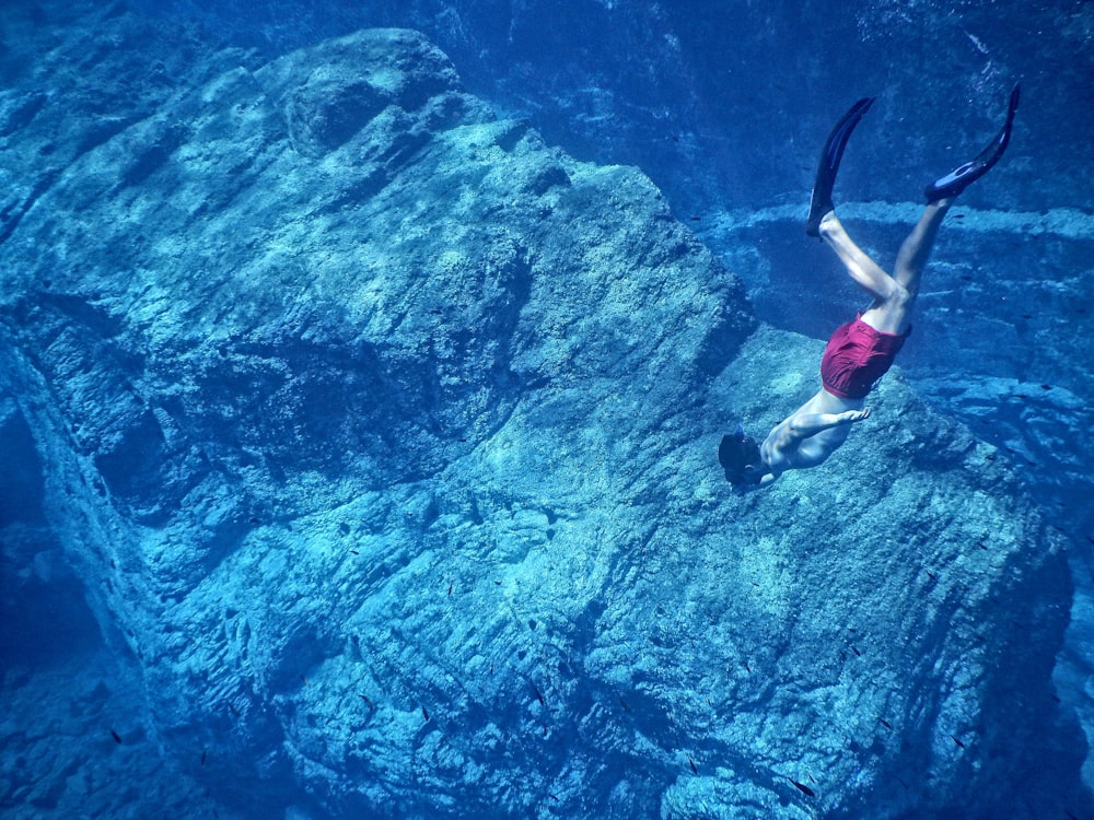 man in red shorts swimming near huge underwater rock