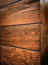 The image displays wooden planks with a textured grain surface, aligned horizontally forming a corner. The wood has a natural, rustic appearance with visible knots and grain patterns.