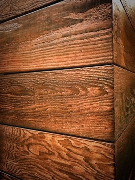 The image displays wooden planks with a textured grain surface, aligned horizontally forming a corner. The wood has a natural, rustic appearance with visible knots and grain patterns.