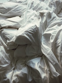 A close-up view of crumpled white bed sheets, creating an abstract pattern with various folds and creases. The fabric appears soft and the lighting casts gentle shadows, highlighting the texture.