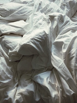 A close-up view of crumpled white bed sheets, creating an abstract pattern with various folds and creases. The fabric appears soft and the lighting casts gentle shadows, highlighting the texture.