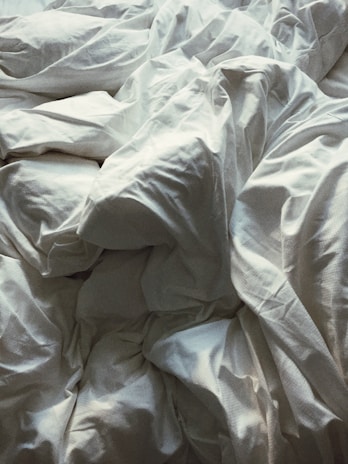 A close-up view of crumpled white bed sheets, creating an abstract pattern with various folds and creases. The fabric appears soft and the lighting casts gentle shadows, highlighting the texture.