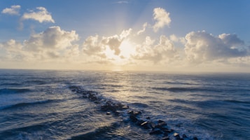 The image captures a serene seascape with the sun rising or setting on the horizon. Fluffy clouds scatter across the sky, and a stone jetty extends into the ocean, dividing the gentle waves.