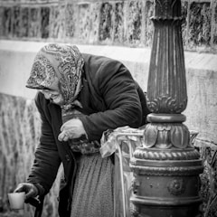 An elderly person with a patterned headscarf and dark overcoat bends over beside a decorative metal post, holding a cup. The scene is in black and white, emphasizing the textures of the clothing and surrounding stone and metal surfaces.