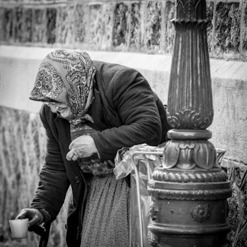 An elderly person with a patterned headscarf and dark overcoat bends over beside a decorative metal post, holding a cup. The scene is in black and white, emphasizing the textures of the clothing and surrounding stone and metal surfaces.