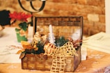 A decorative wooden basket filled with festive items such as a small figurine, greenery, orange leaves, and a candle, set against a brick wall. A vase with a red poinsettia is in the background, and the scene is lit with warm lighting.