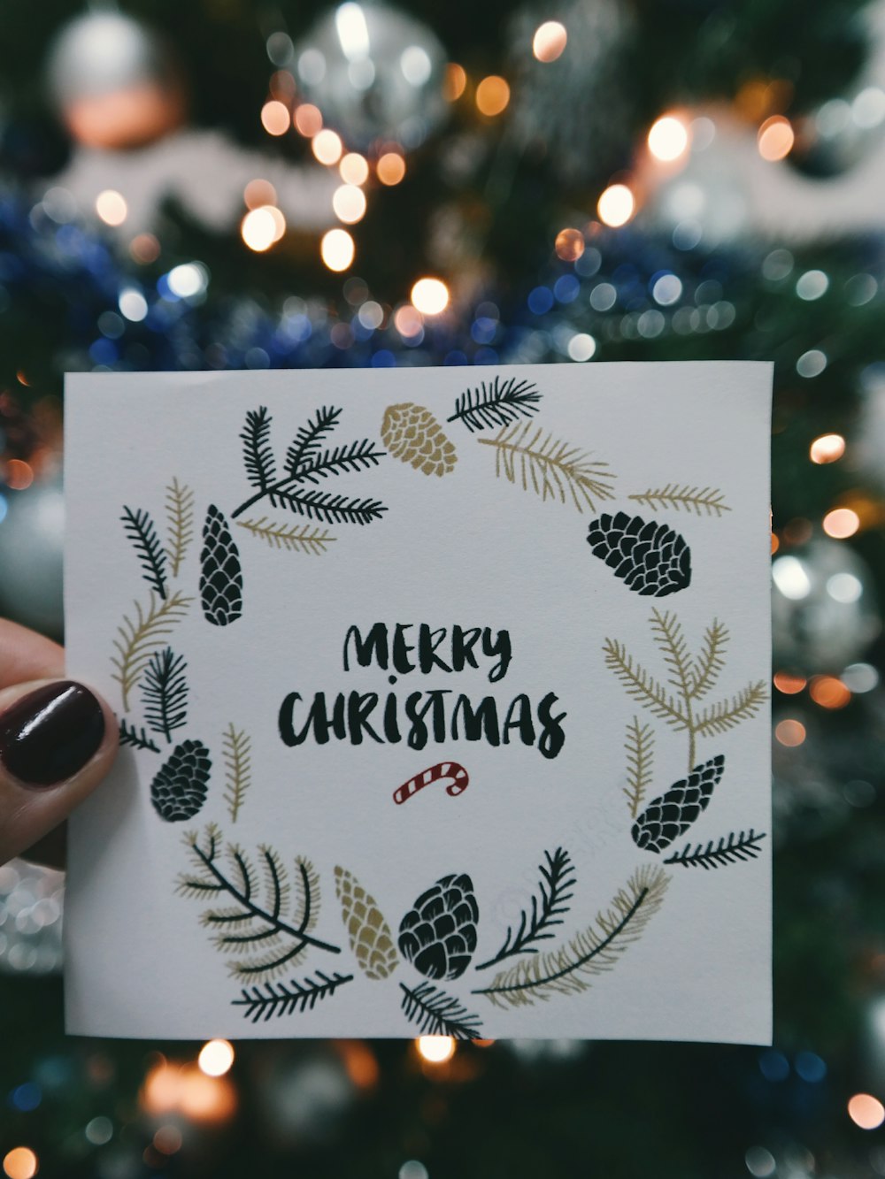 Ein Stück Papier mit der Aufschrift "Frohe Weihnachten" mit einem Kranz drumherum, das vor einem Weihnachtsbaum hochgehalten wird.