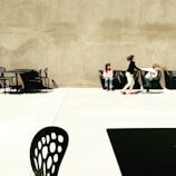 Three children are interacting in an outdoor, minimalist space with modern, black furniture. One child sits alone while two others playfully engage with each other. The area is spacious, with a stark contrast between the light concrete ground and the dark chairs and tables.