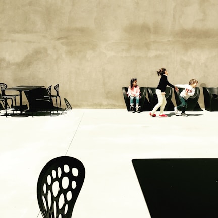 Three children are interacting in an outdoor, minimalist space with modern, black furniture. One child sits alone while two others playfully engage with each other. The area is spacious, with a stark contrast between the light concrete ground and the dark chairs and tables.