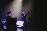 A stage performance with a musician holding up one hand, standing in front of a microphone. There are musical instruments and equipment visible, including a keyboard and a music stand. The scene is illuminated by soft stage lighting, highlighting the performer's presence.