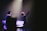 A stage performance with a musician holding up one hand, standing in front of a microphone. There are musical instruments and equipment visible, including a keyboard and a music stand. The scene is illuminated by soft stage lighting, highlighting the performer's presence.