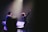 A stage performance with a musician holding up one hand, standing in front of a microphone. There are musical instruments and equipment visible, including a keyboard and a music stand. The scene is illuminated by soft stage lighting, highlighting the performer's presence.
