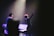 A stage performance with a musician holding up one hand, standing in front of a microphone. There are musical instruments and equipment visible, including a keyboard and a music stand. The scene is illuminated by soft stage lighting, highlighting the performer's presence.