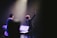 A stage performance with a musician holding up one hand, standing in front of a microphone. There are musical instruments and equipment visible, including a keyboard and a music stand. The scene is illuminated by soft stage lighting, highlighting the performer's presence.