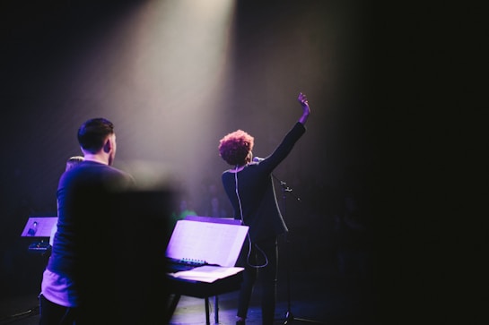 A stage performance with a musician holding up one hand, standing in front of a microphone. There are musical instruments and equipment visible, including a keyboard and a music stand. The scene is illuminated by soft stage lighting, highlighting the performer's presence.