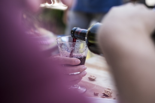 A person is pouring red wine into a glass with an intricate floral design. The scene includes a hand holding the glass and a blurred foreground, creating a casual and intimate setting. Crumbs or pieces of food are scattered on a wooden table, suggesting a relaxed dining or social gathering.