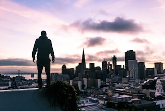 A silhouette of a person stands on a rooftop, overlooking a cityscape during what appears to be either sunrise or sunset. Prominent skyscrapers, including the distinct pyramid-shaped Transamerica Pyramid, are visible. Dark clouds hover over the city while the sky is tinged with soft hues of pink, purple, and orange. The distant bridge and various buildings contribute to the urban scene.