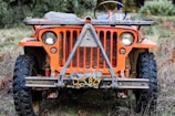 A frontal view of a vintage, weathered orange jeep with a California license plate, parked on a grassy terrain. The jeep's design is rugged with noticeable rust and signs of age, featuring round headlights and large off-road tires.