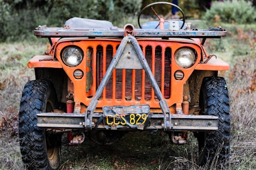 A frontal view of a vintage, weathered orange jeep with a California license plate, parked on a grassy terrain. The jeep's design is rugged with noticeable rust and signs of age, featuring round headlights and large off-road tires.