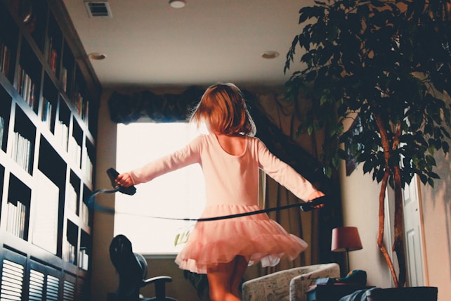 A child in a pink dress twirls around a room filled with bookshelves on the left and a large potted plant on the right. The room is softly lit with natural light coming from a window behind the child.