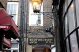 A narrow alleyway between brick buildings, featuring a classic street lamp. A sign above an archway reads 'Bedfordbury No. 24 No. 23'. On the left, a red awning with white text reads 'Giovanni's'. Large windows with multiple panes are visible on the right side.