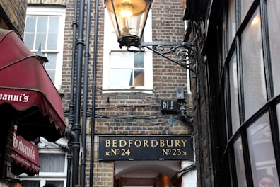 A narrow alleyway between brick buildings, featuring a classic street lamp. A sign above an archway reads 'Bedfordbury No. 24 No. 23'. On the left, a red awning with white text reads 'Giovanni's'. Large windows with multiple panes are visible on the right side.