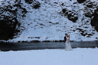 A bride and groom stand closely together near a riverbank, surrounded by a snowy landscape. The groom is in a black suit while the bride wears a white wedding dress. Snow covers the ground and rocky cliffs in the background.