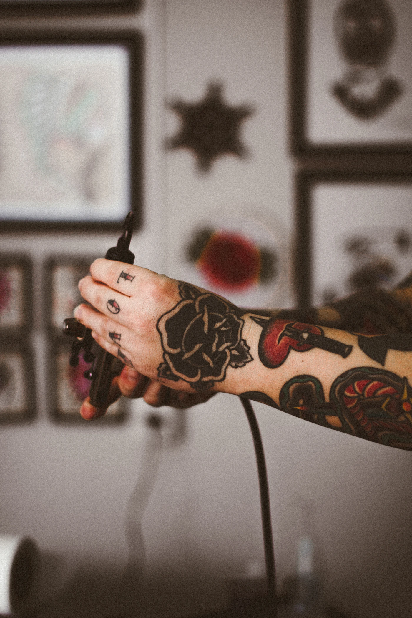 Close-up of a tattooed hand holding a black tattoo machine, with framed artwork blurred in the background.
