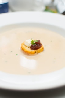A creamy soup is presented in a wide, white soup bowl. Floating in the center is a round crouton topped with a dollop of white cream or cheese, a small amount of dark red jam or sauce, and a sprig of fresh green herb.