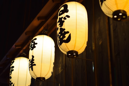 A row of illuminated Japanese paper lanterns with black kanji characters hanging from a wooden structure. The background features a dimly lit wooden wall, enhancing the warm glow of the lanterns.