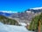 A picturesque alpine scene featuring a snow-covered ski slope descending into a charming mountain village. Dense evergreen forests frame the view on either side, while majestic, snow-capped peaks rise in the background against a clear blue sky.