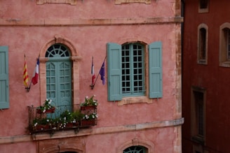 A rustic building facade painted in a warm pink hue with blue-green shutters and a matching door. Three flags, including the French flag and possibly regional or European flags, are mounted on the wall next to a small balcony adorned with pots of white flowers and greenery.