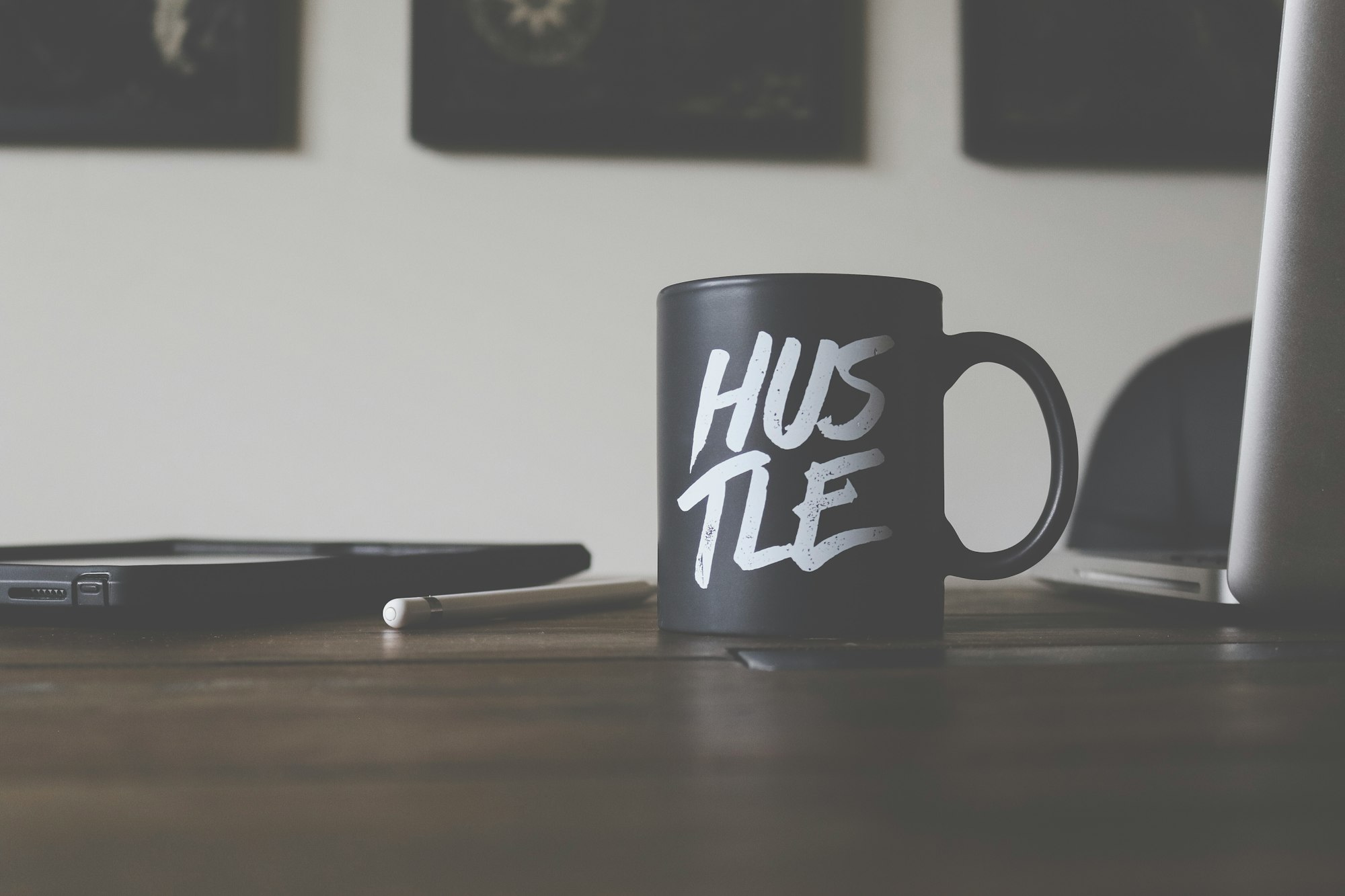 a black coffee mug reading "hustle" next to an open laptop and a pad with a stylus, all on a woodes desk