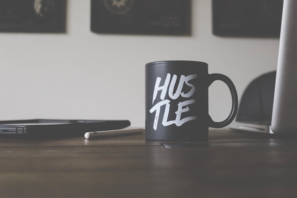 a black coffee mug reading "hustle" next to an open laptop and a pad with a stylus, all on a woodes desk