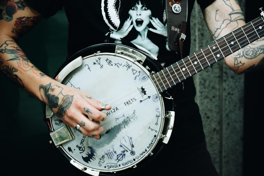 A person with tattooed arms is playing a banjo adorned with various drawings and writings. The individual is wearing a black T-shirt featuring a surprised figure. The scene is likely set against an urban backdrop.