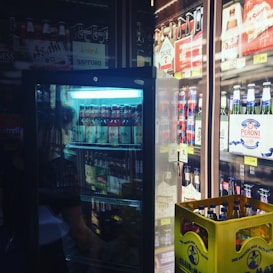 A refrigerated display filled with various beer bottles and cans. The fridge is partially open, revealing brands like Sapporo, Asahi, and Peroni. There is a yellow crate filled with bottled club sodas or similar beverages on the floor. Bright lighting highlights the labels and colors of the products.