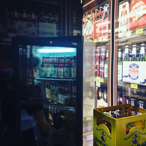 A refrigerated display filled with various beer bottles and cans. The fridge is partially open, revealing brands like Sapporo, Asahi, and Peroni. There is a yellow crate filled with bottled club sodas or similar beverages on the floor. Bright lighting highlights the labels and colors of the products.