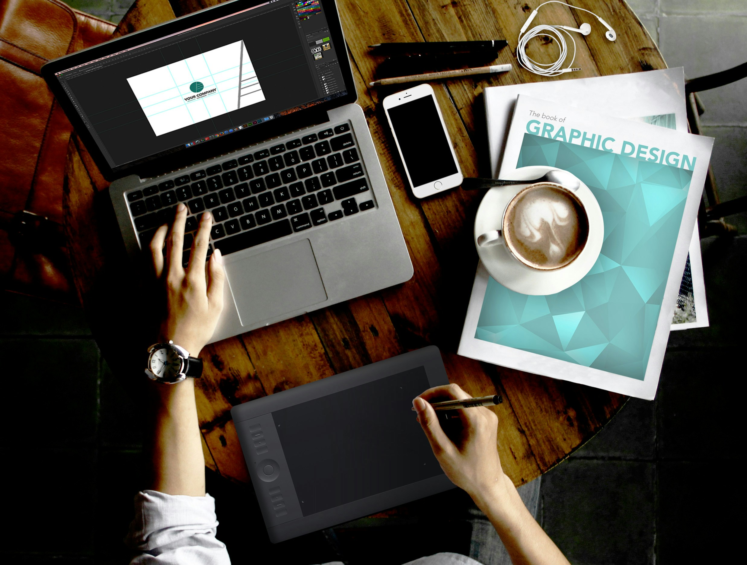 A person is working on a wooden table with various items. A laptop displays a design program, while the person uses a graphic tablet with a stylus. Nearby are a magazine titled 'Graphic Design', a smartphone, a cup of coffee, and a pair of earphones. The watch on the person's wrist suggests attention to time management.