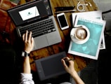 A person is working on a wooden table with various items. A laptop displays a design program, while the person uses a graphic tablet with a stylus. Nearby are a magazine titled 'Graphic Design', a smartphone, a cup of coffee, and a pair of earphones. The watch on the person's wrist suggests attention to time management.