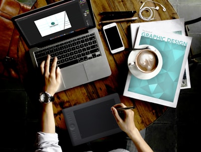 A person is working on a wooden table with various items. A laptop displays a design program, while the person uses a graphic tablet with a stylus. Nearby are a magazine titled 'Graphic Design', a smartphone, a cup of coffee, and a pair of earphones. The watch on the person's wrist suggests attention to time management.