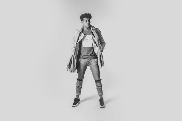 A person poses confidently in a studio setting with a neutral background, wearing layered casual clothing including a coat and scarf. The individual stands with one foot slightly forward, exuding a sense of style and self-assurance.