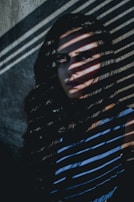 A person is partially obscured by dramatic shadow patterns cast across their face and body by blinds. The image features strong contrast between light and shadow, with the person’s expression appearing intense and mysterious. The background appears to be a plain, textured wall.