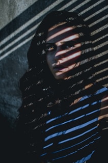 A person is partially obscured by dramatic shadow patterns cast across their face and body by blinds. The image features strong contrast between light and shadow, with the person’s expression appearing intense and mysterious. The background appears to be a plain, textured wall.