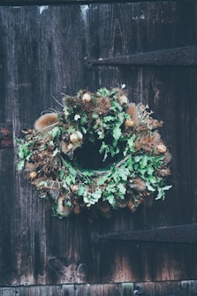 A wreath made of dried leaves, thistles, and other natural materials hangs on a rustic wooden door with visible grain and an old metal hinge.